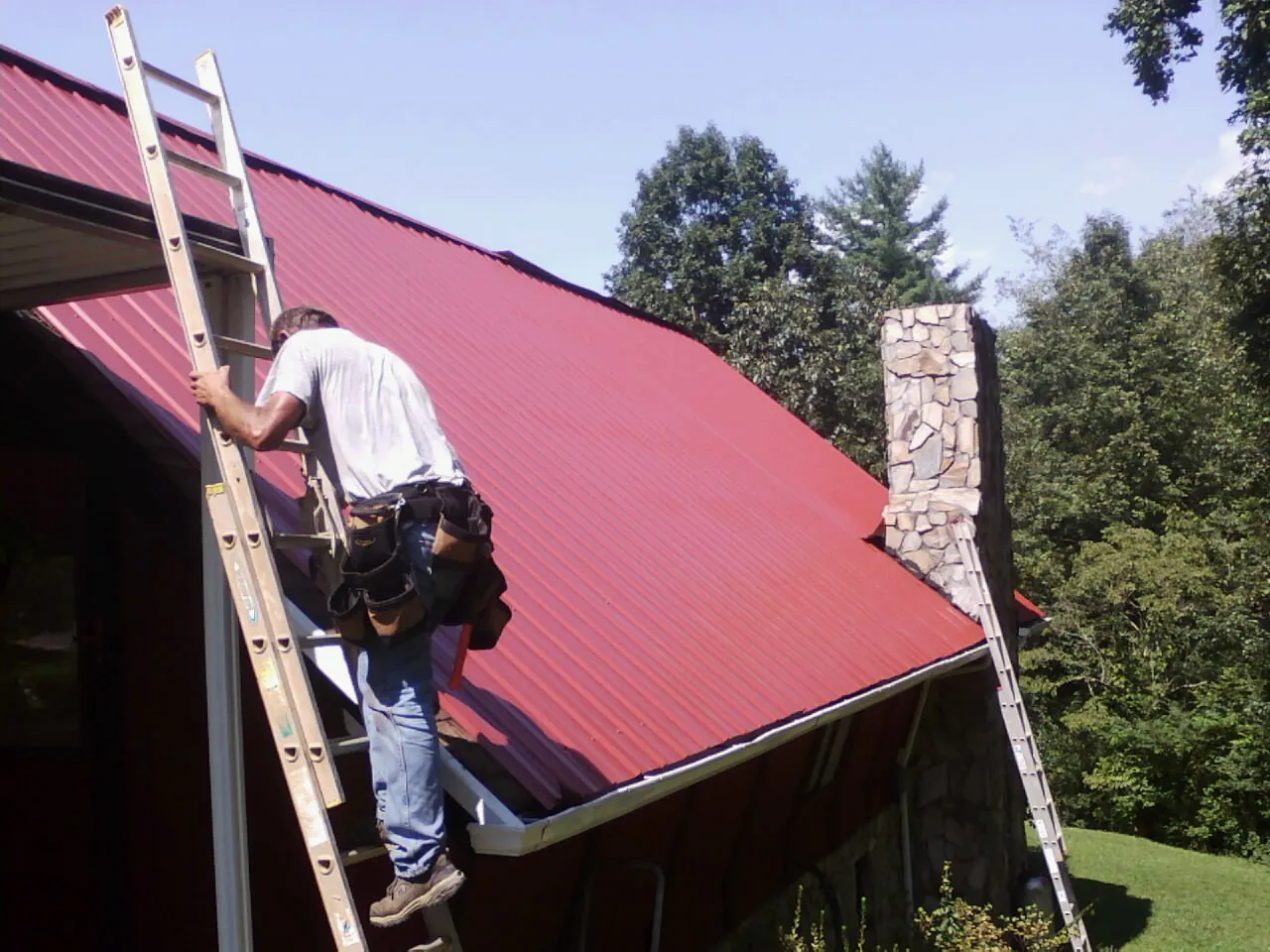 Roofer on a ladder beside a red metal roof and stone chimney — Soffit & Fascia Repair by Roofing Brothers in Delaware City